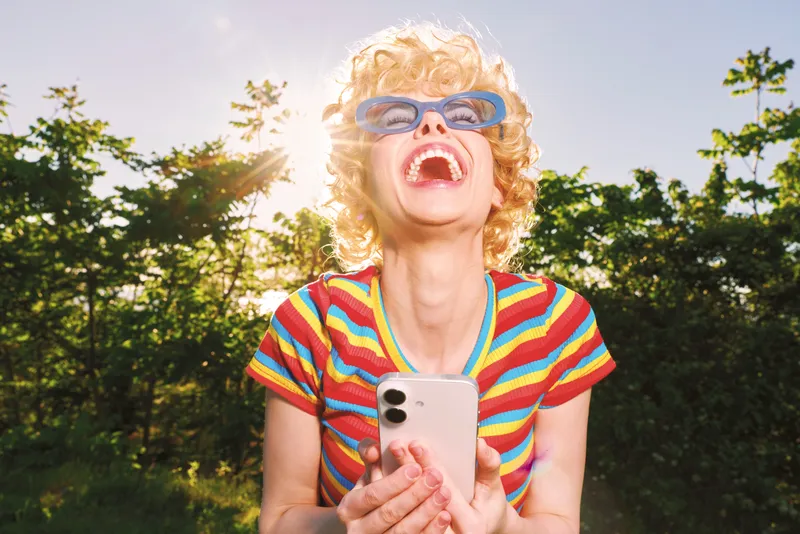 Vibrant outdoor portrait of a joyful woman wearing a striped colorful shirt and playful blue sunglasses, holding a smartphone, captured with retro film mood and bright sunlight, sharp focus, sunlit