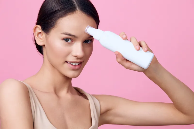 Young woman showcasing a beauty product on her forehead with a cheerful smile, set against a soft pink background, reflecting a fresh and vibrant skincare concept
