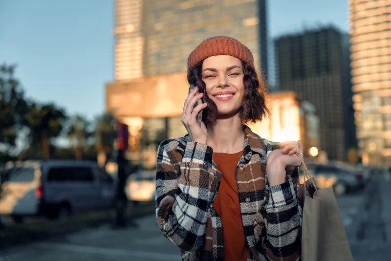 Woman smiling on phone while shopping in city during golden hour, candid lifestyle moment with authenticity and warm glow; beanie and plaid jacket convey modern mindful urban joy.
