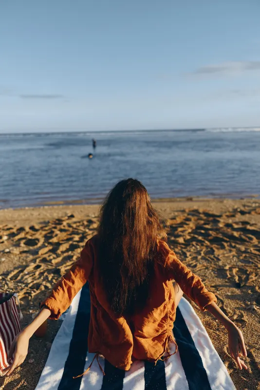 Woman on beach, back to camera, long hair catching sunlight, sunset reflections across sea horizon, relaxed mood, sand texture underfoot, gentle waves and clear sky