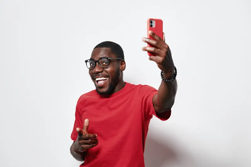 African American man with dark skin wearing glasses and red shirt taking selfie with smartphone, smiling and posing against white background in casual style and happy mood.