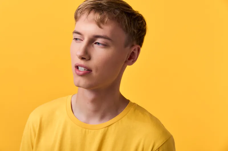 Young man wearing a yellow shirt stands before a vibrant yellow backdrop, expressing casual style, contemplative mood and modern fashion suitable for lifestyle and brand projects.