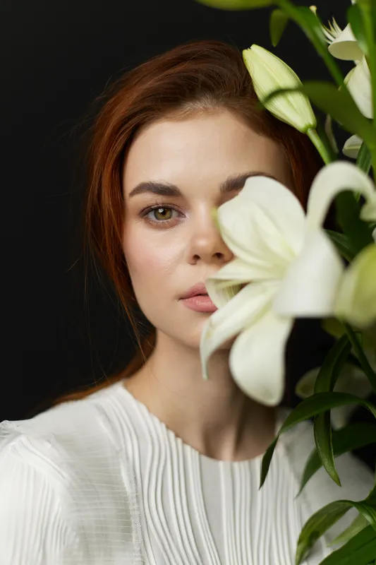 A beautiful young woman with long auburn hair peeking through white lilies, showcasing a serene expression against a dark background, highlighting natural beauty and elegance