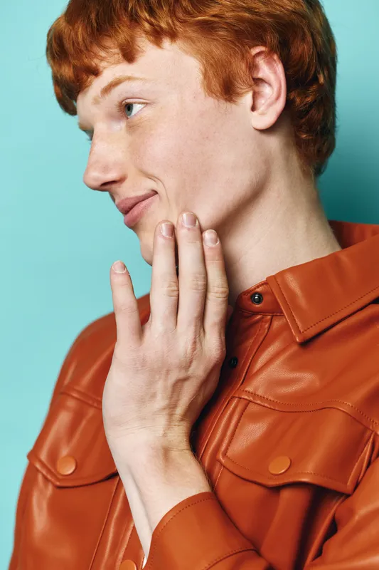 Young man with red hair wearing orange jacket touching face and looking sideways with calm expression against blue background in studio portrait with soft lighting.