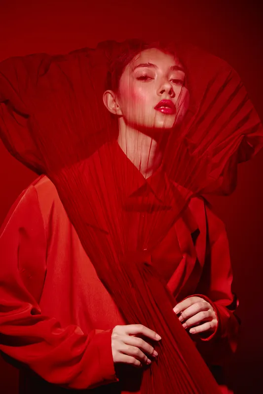 Fashion model poses dramatically in bold red outfit enhanced with sheer fabric animation. She emphasizes high-end beauty with intense makeup. Studio artistic concept in vibrant red tone.