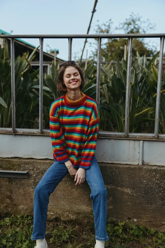 Woman smile sweater jeans sitting outdoor portrait casual young adult wearing colorful striped knit sweater and denim, relaxed pose on low wall with greenery background.