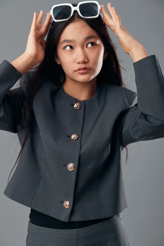 Young woman wearing gray jacket adjusting white sunglasses on head looking sideways with long dark hair and minimal makeup on gray background in studio portrait.