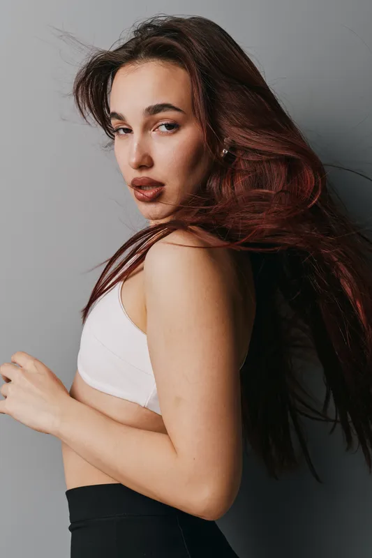Young woman with long brown hair and natural makeup posing in white top and black pants against gray background. Beauty, fashion and confident model with casual style and expression.
