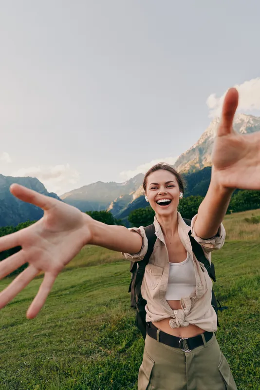 Young woman with a backpack rushes toward the camera with outstretched arms in a bright mountain meadow, smiling and energized by the outdoor space.