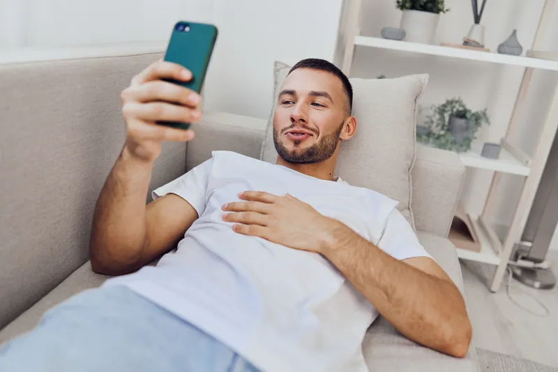 Relaxed man lounging at home, engaging with his smartphone while embracing digital detox and solo living decisions. A moment of simplicity and authenticity.