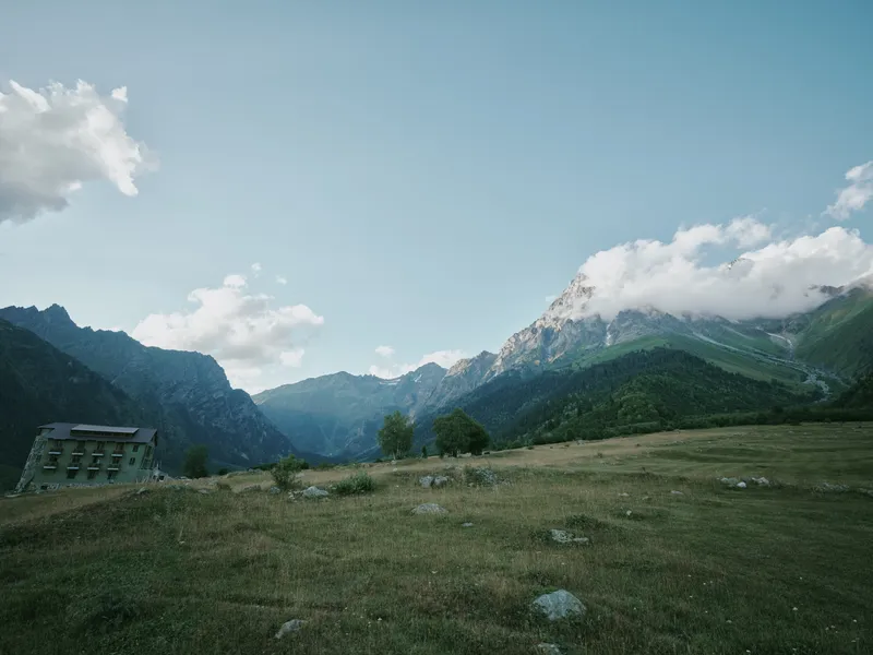 Mountain landscape, meadow, building, valley, clouds, sky, rural, grass, nature, scenery, expansive hillside view with distant peaks and a lone structure for exploration and photography adventure