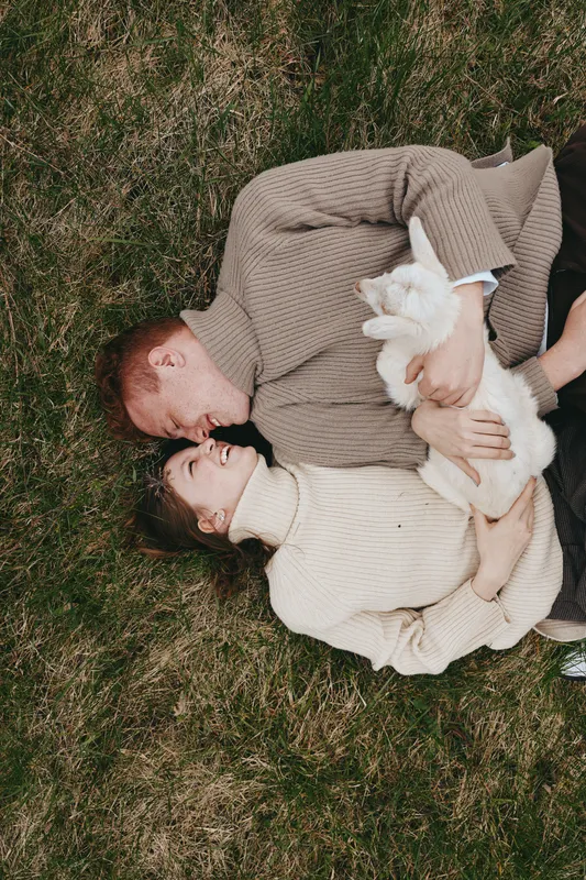 Young couple lying on grass outdoors holding a white baby goat while smiling and enjoying a cozy autumn day together in warm knitted sweaters.