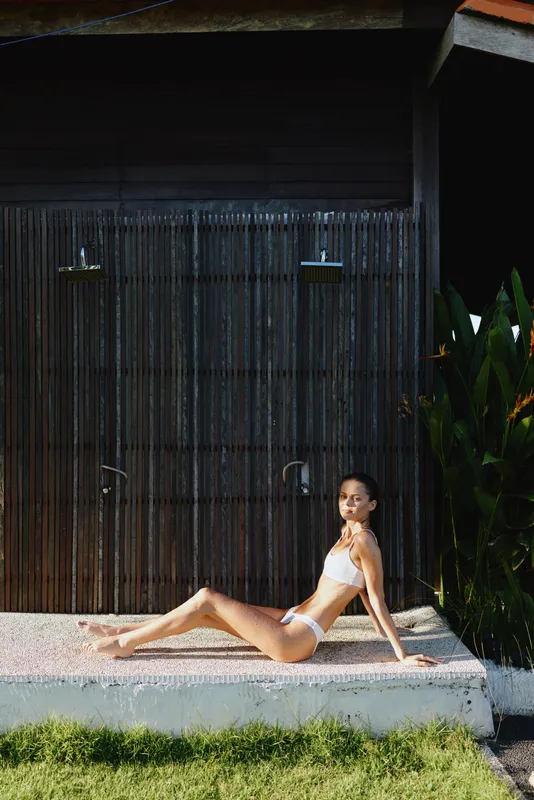 woman, bikini, outdoor, summer, sun, relaxation, model, poolside, fashion, lifestyle. A serene scene of a woman in a white bikini lounging on a sunlit edge beside a dark wall, evoking casual vacation
