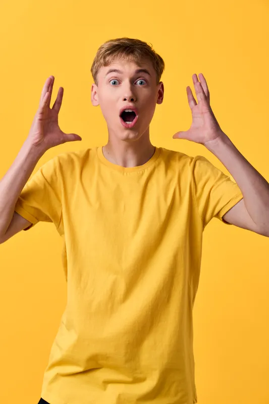 Surprised man in a bright yellow shirt shows open-mouthed shock with raised hands, capturing a playful moment against a vivid background