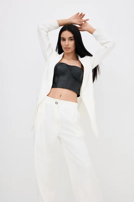 Young woman wearing stylish white suit jacket and pants with black leather corset top, posing confidently against a plain white background in modern fashion style.