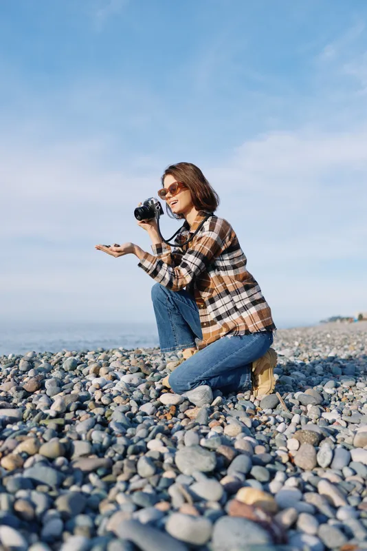 Photographer, woman on a rocky beach kneels with camera, capturing outdoor nature and travel mood in casual style under a bright sky with calm water in the background