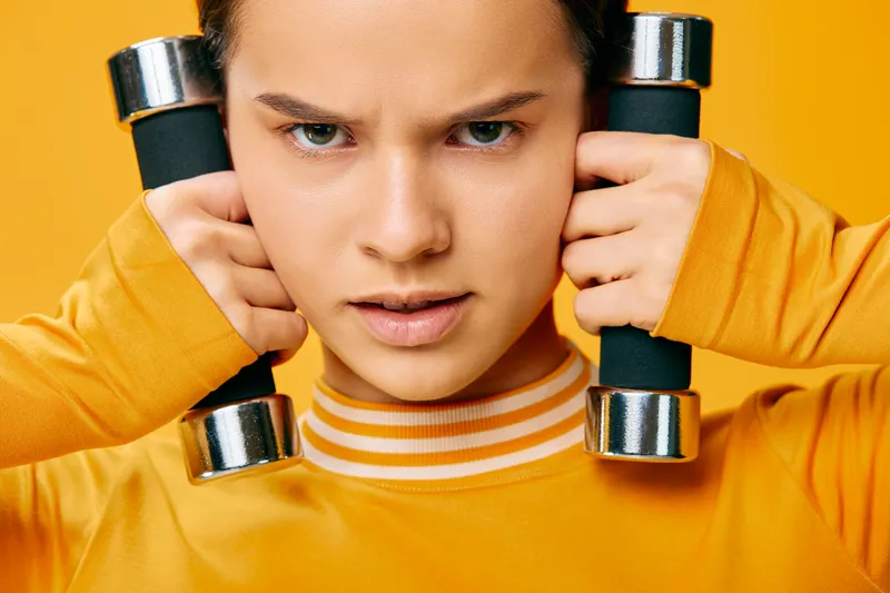 Close-up of a focused woman in a bright yellow athletic top holding dumbbells at her shoulders, ready for a vigorous workout. Bold color background highlights fitness energy and athletic