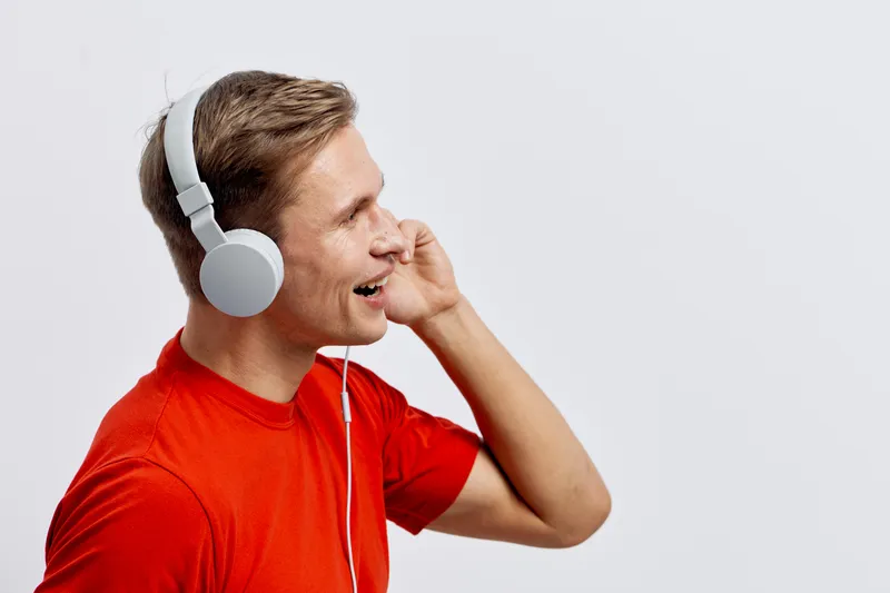 Smiling young man with headphones listening to music isolated on plain light background. Casual clothing, cheerful expression, modern lifestyle, leisure activity, happy mood, male model.