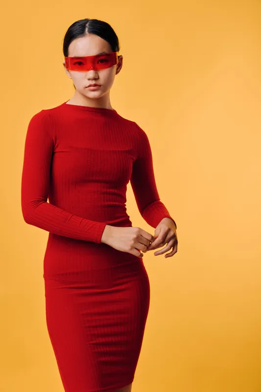 Woman in red fitted dress wearing red transparent visor glasses standing against orange background with serious expression and elegant posture for fashion and style.