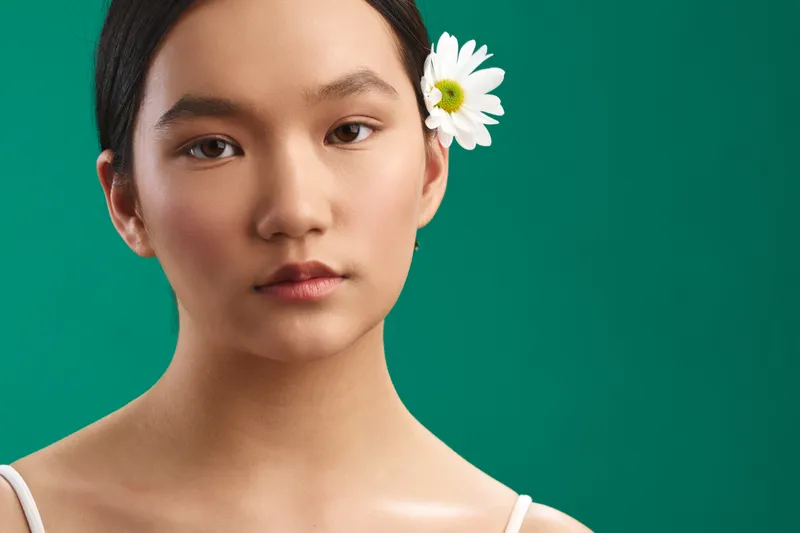 asian woman portrait closeup featuring a calm face with a white flower tucked behind her ear, set against a vivid green background.