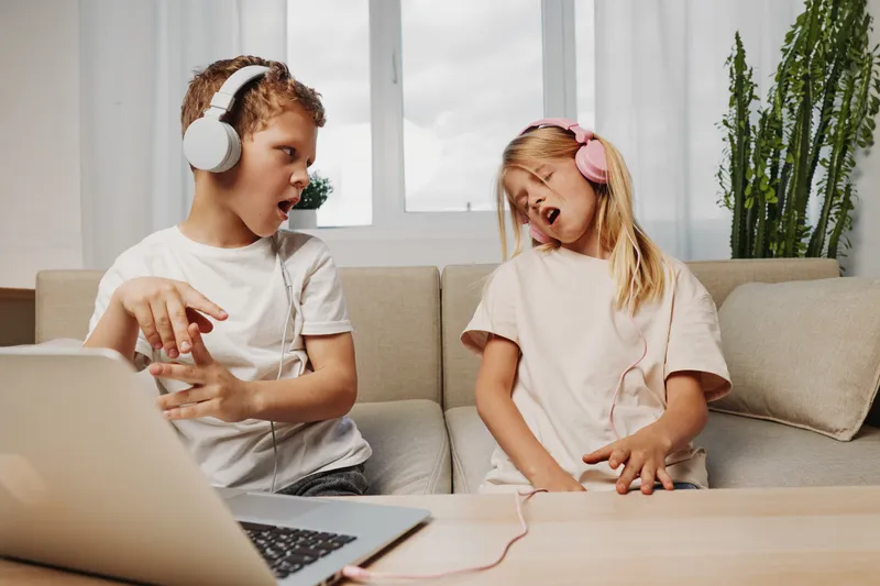 Two kids with headphones play air guitar on couch near laptop in bright room, enjoying music and fun together. Concept for childhood, entertainment, and technology.