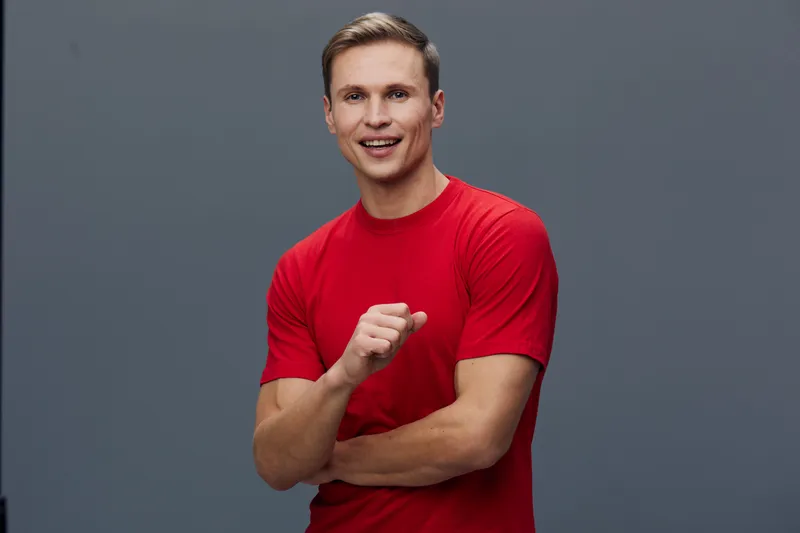 Cheerful young man wearing red t-shirt smiling and pointing at himself on plain gray background. Happy, confident, casual, casualwear, male, middle-aged, positive vibe, studio portrait, people lifesty