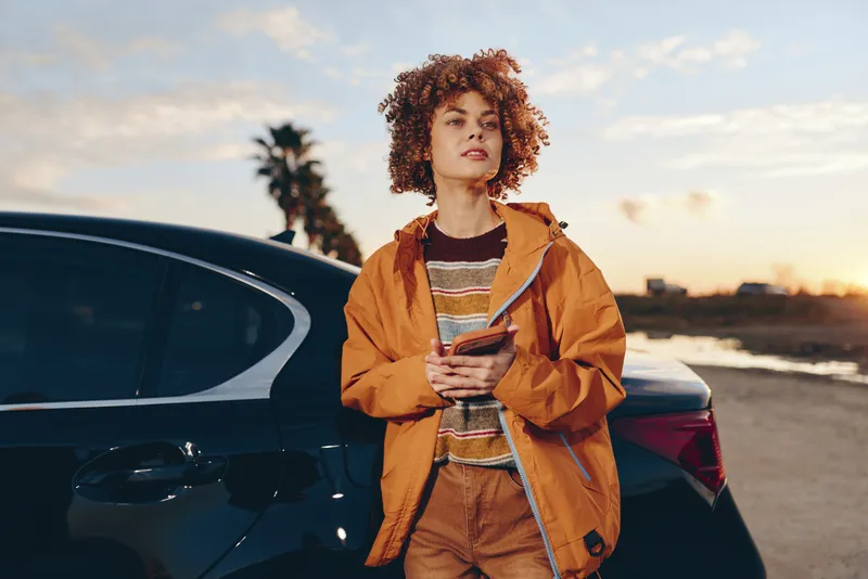Woman lifestyle outdoor by car at sunset, wearing orange jacket and striped top, curly hair, holding phone and smiling calmly. Evening road background suggests travel and communication.