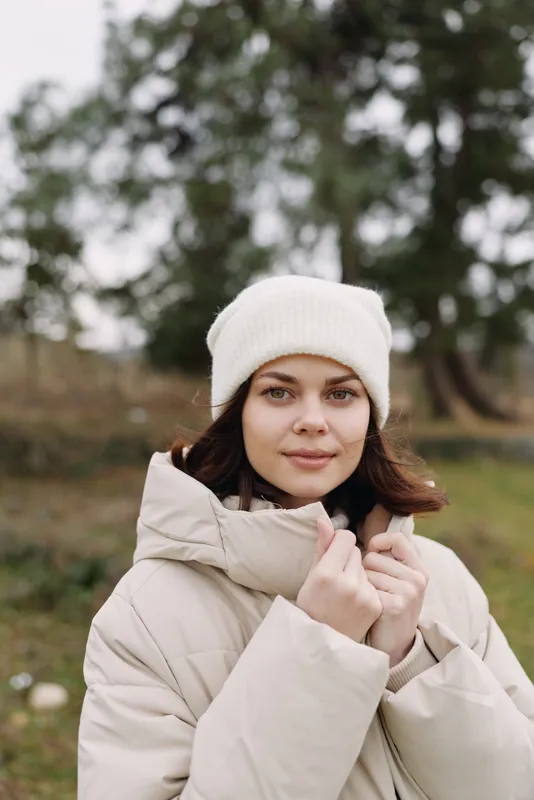 Woman wearing a beige puffer coat and white beanie outdoors standing in a misty park looking at camera with soft natural light and calm mood