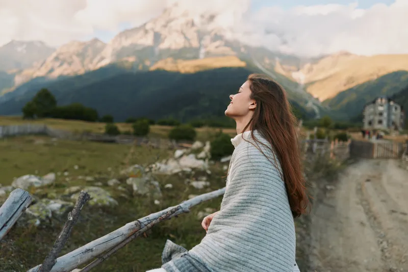 A woman stands along a country road wrapped in a blanket, gazing at distant mountains under a clear sky, conveying calm, solo travel and reflective lifestyle.