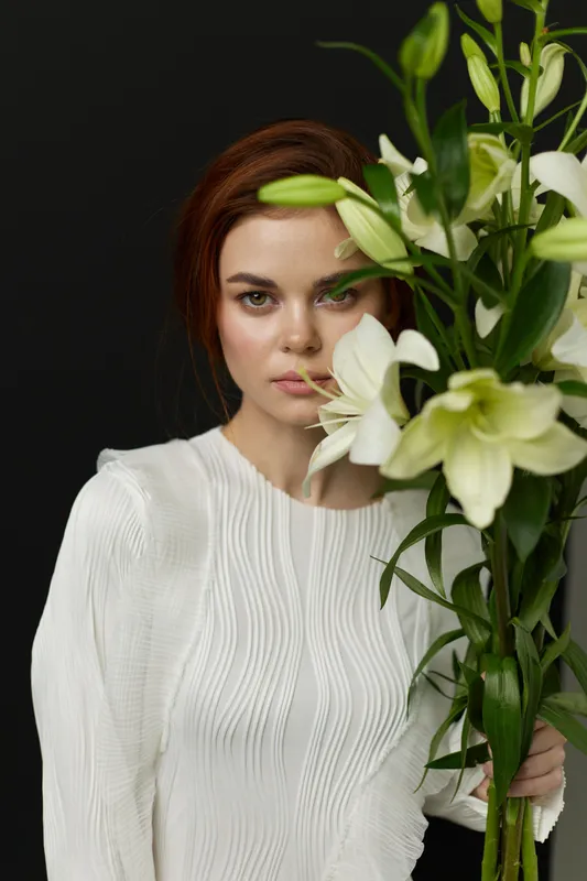 Female model with long hair holds white lilies, wearing a textured white blouse against a dark background, conveying elegance and serenity