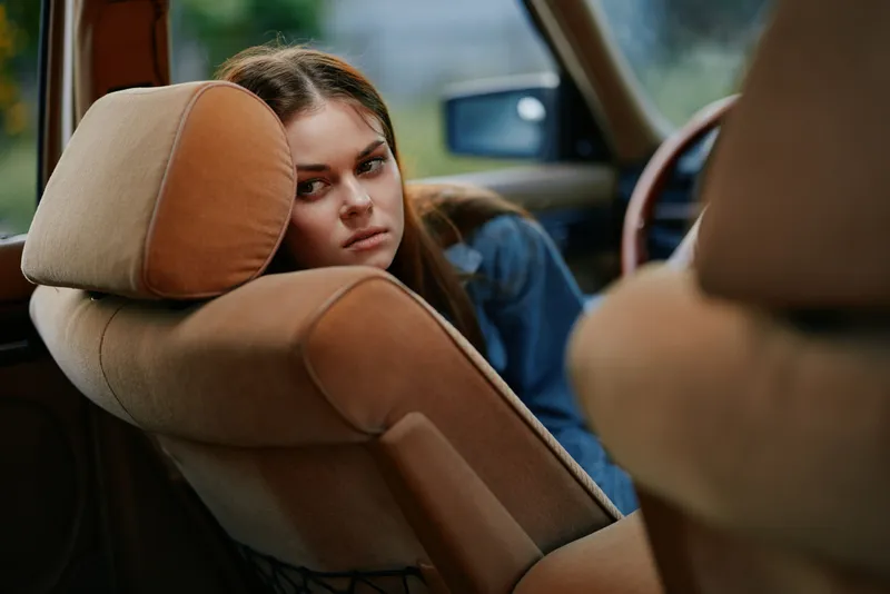 Young woman with sad expression sitting in a vintage car, dressed in casual attire, reflecting on life moments in a nostalgic atmosphere.