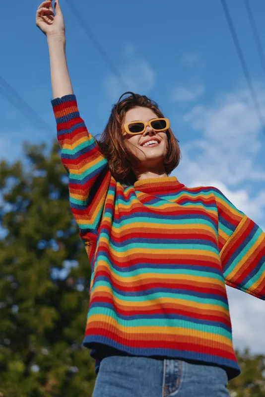 woman in rainbow sweater and sunglasses smiling outdoors, young adult with raised arm in casual outfit against blue sky. Lifestyle travel vibe, joyful expression and retro street fashion