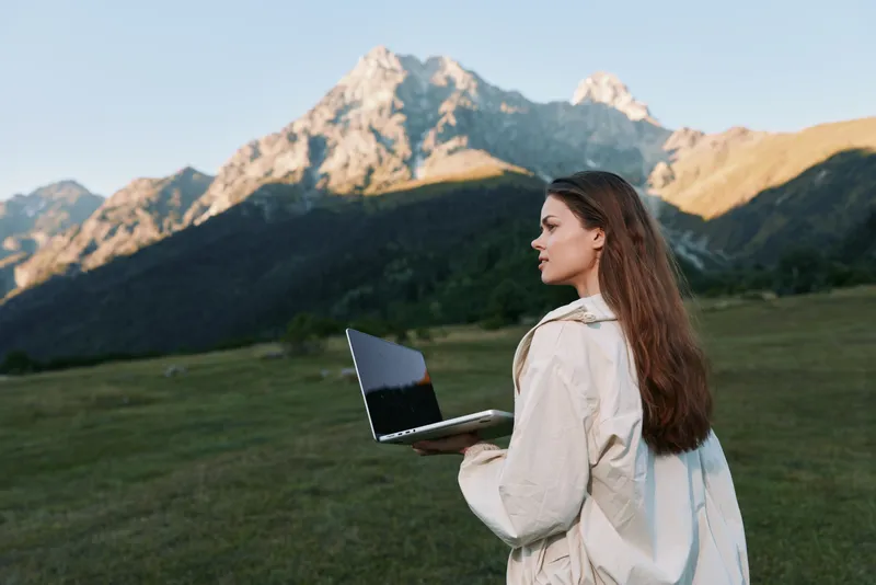 A focused woman stands outdoors in a grassy field with a laptop, gazing into the distance as sunlight highlights the mountains behind her, creating an adventurous work scene.
