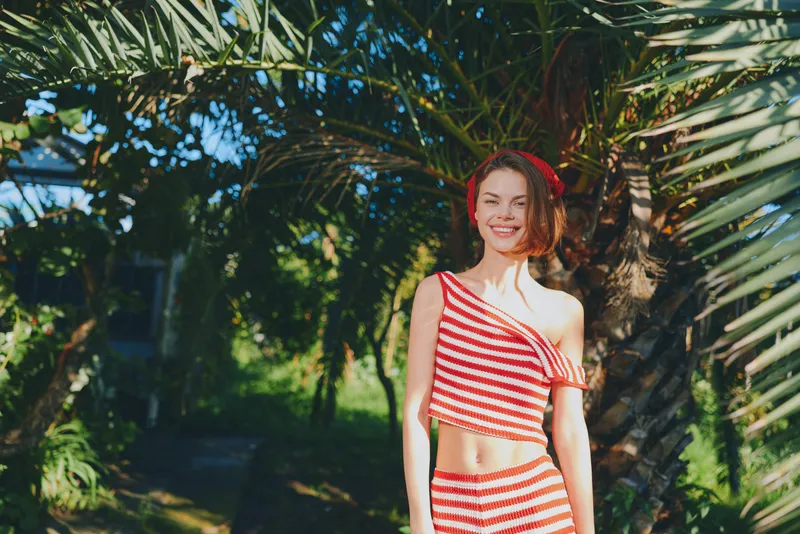 A cheerful woman in a red and white striped outfit stands in a sunny tropical garden. She smiles warmly, posing amid palm trees, creating a lively, carefree vibe.