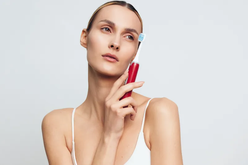 A young woman with clear skin holds a red electric toothbrush near her face, looking thoughtful against a plain white background. Concept for dental care and personal hygiene.