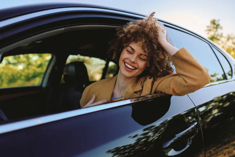 Smiling woman with curly hair leaning out of a car window on a sunny day. Casual fashion vibe with retro film color tones, capturing a warm, candid lifestyle moment.