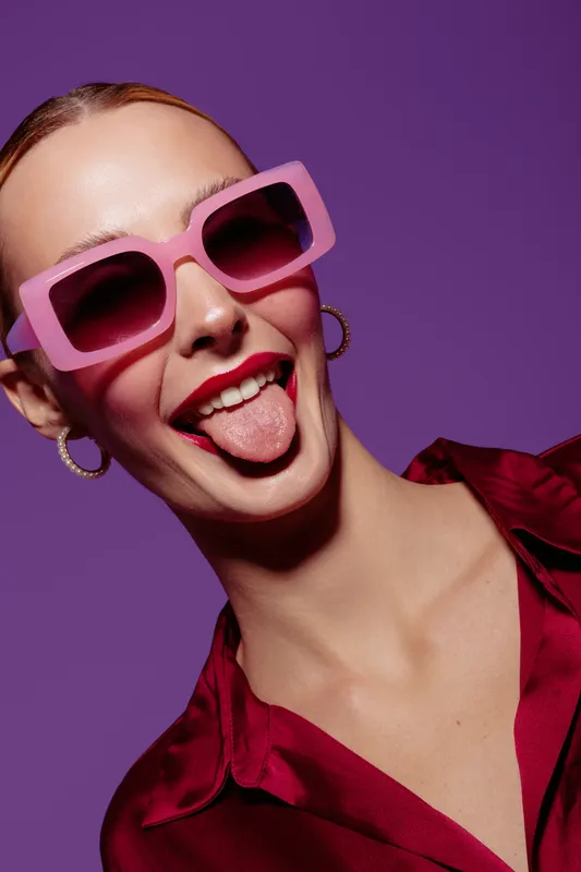 Young woman with pink sunglasses and red lipstick sticks out tongue on purple background. Fashion portrait with playful expression and trendy style in studio lighting.
