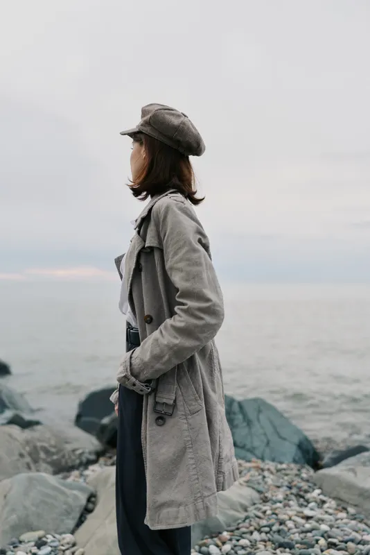 Woman in warm coat and hat stands on rocky beach looking at gray sea under overcast sky. Autumn or winter mood with casual fashion and calm ocean background. Concept for solitude and nature.