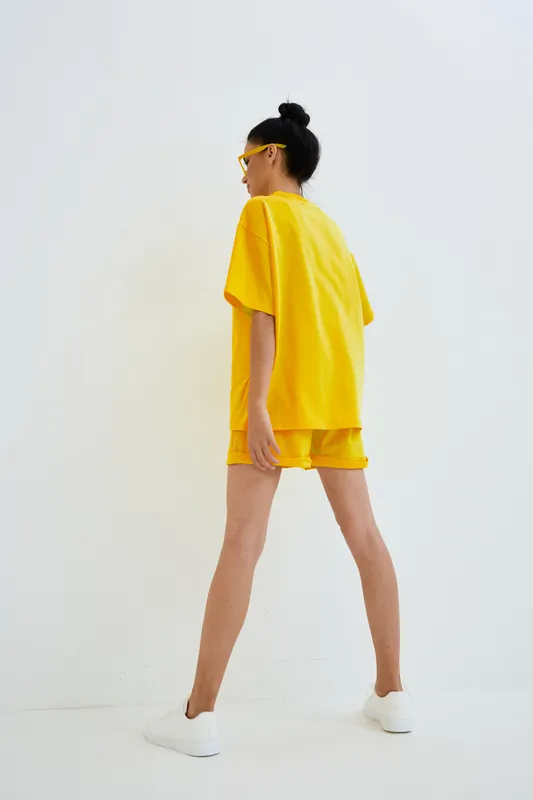 Young woman wearing bright yellow oversized t-shirt and shorts stands confidently against a plain white background, showcasing casual summer style with sporty white sneakers.