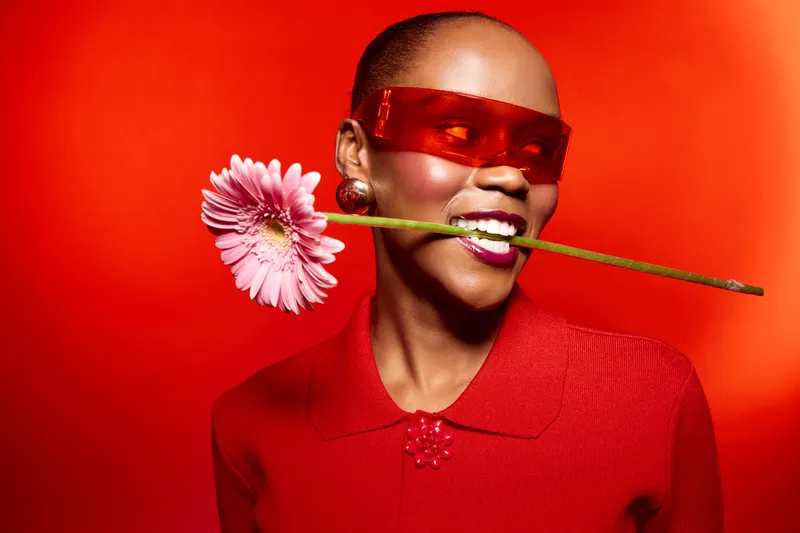 Smiling black woman with dark skin holds pink flower in mouth wears red sunglasses and red top on bright red background. Concept of beauty, fashion, and bold colorful style portrait.