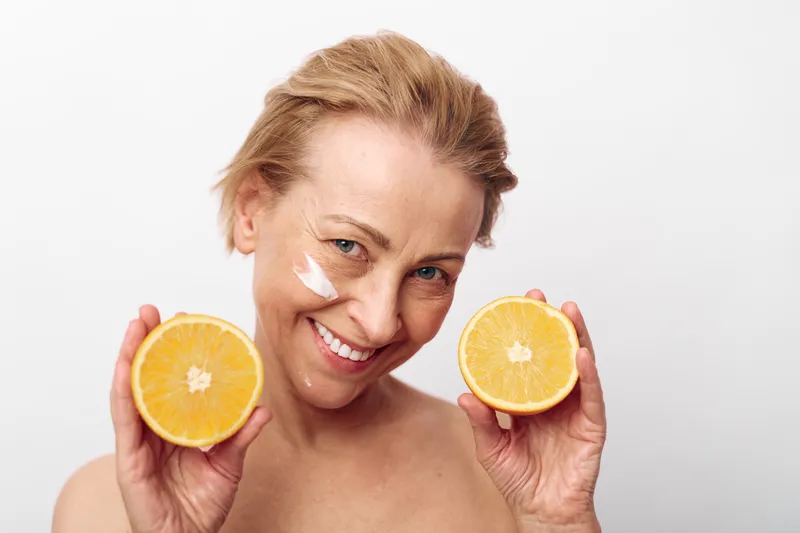 Smiling middle aged woman holds two orange halves near face with facial cream on cheek, isolated on white background. Concept of natural skincare, healthy lifestyle, and citrus beauty treatment.