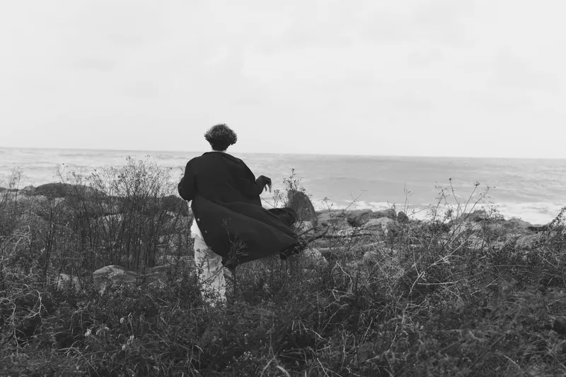 Solitude seascape on a windy cliff with a lone silhouette overlooking the ocean, sparse vegetation and expansive horizon, monochrome mood conveying contemplation and calm.