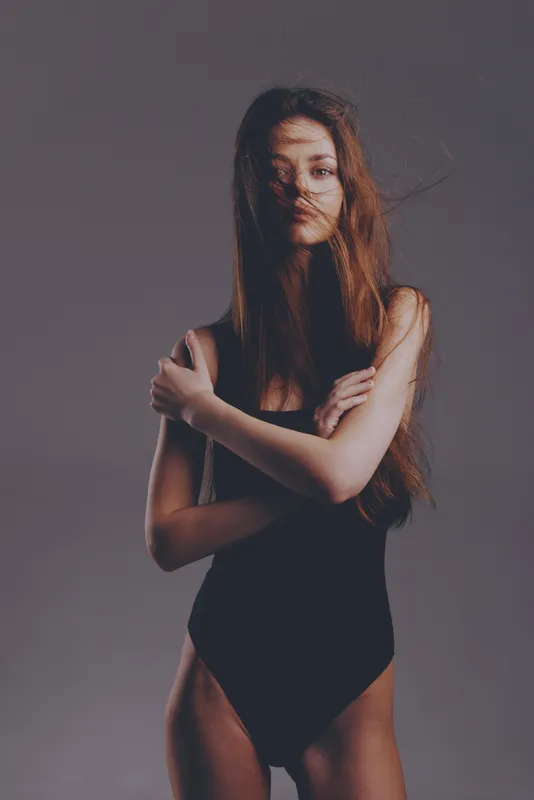 young woman in black bodysuit posing with crossed arms in studio with moody lighting