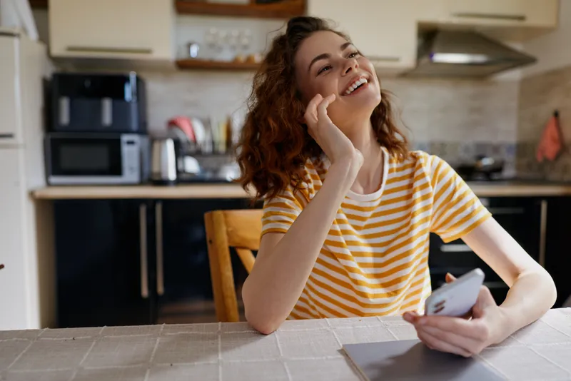 Young woman with curly hair chats on a smartphone at a kitchen table, smiling as she enjoys a light, casual conversation in a bright, modern home setting, creating a welcoming mood.