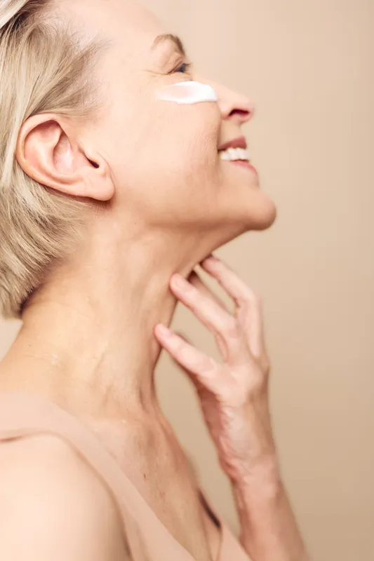 Smiling mature woman applies white face cream on cheek, showing skincare routine and self-care. Close-up portrait with soft lighting on neutral background, concept of beauty and wellness.