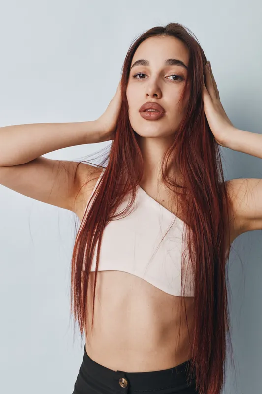 Young woman with long straight hair posing with hands near head against white background. Fashion model wearing crop top and black pants in studio with confident expression and natural makeup.