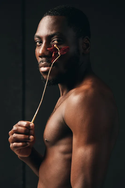 Black man with dark skin holding dried flower near face in creative conceptual visual style. Portrait of African ethnicity with bare torso and artistic expression on dark background.