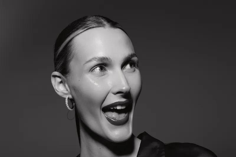 Caucasian woman with light skin and slicked back hair smiles with open mouth looking up. Black and white portrait with high contrast lighting and dark background.