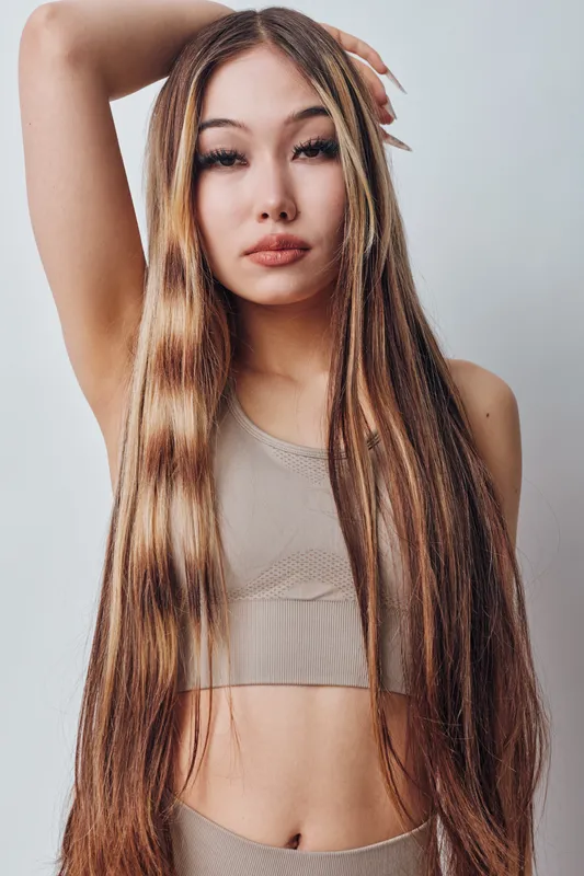 Young woman with long brown hair and neutral makeup wearing beige top posing with arm raised on light background. Beauty, fashion and portrait concept.