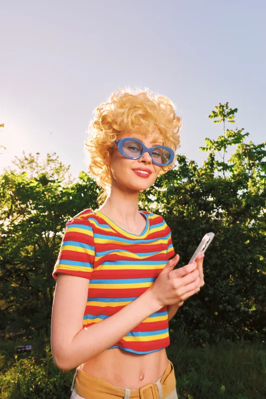 Young woman with a curly blonde wig and rainbow striped top stands outdoors, posing with a phone, retro film color palette, sunny park background, confident lifestyle portrait.